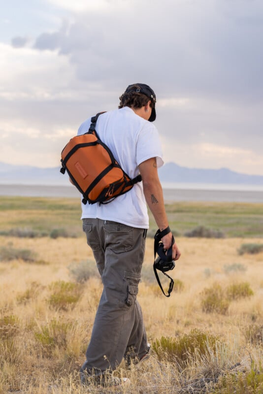A person wearing a white t-shirt, cargo pants, and a black cap walks through a dry grassy field, carrying an orange backpack and holding a camera, with mountains and a cloudy sky in the background.