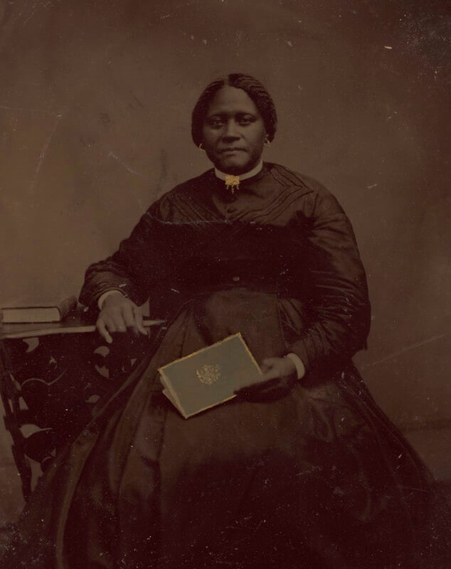 A Black woman in a dark Victorian dress sits beside a table with books, holding a closed book in her lap and looking calmly at the camera. The image is a sepia-toned, historical portrait.