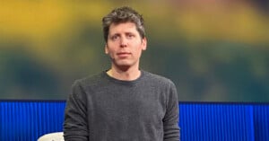 A man with short, tousled brown hair wearing a gray sweater sits in front of a blurred background with blue and yellow tones.