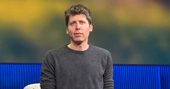 A man with short, tousled brown hair wearing a gray sweater sits in front of a blurred background with blue and yellow tones.