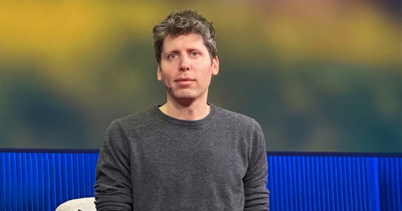 A man with short, tousled brown hair wearing a gray sweater sits in front of a blurred background with blue and yellow tones.