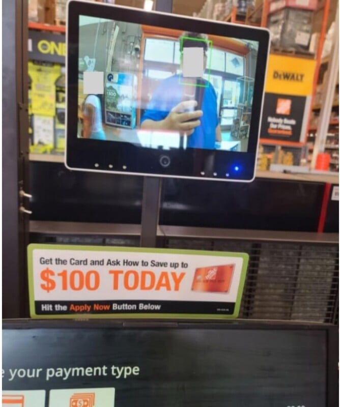 A person is taking a photo of a self-checkout screen at a store, showing their reflection on a security monitor above a sign advertising a credit card offer to save up to $100 at The Home Depot.