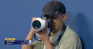 A photographer wearing a cap and green shirt takes a picture with a large camera at a tennis match. The scoreboard shows Medvedev vs. Bonzi, with Bonzi at match point.