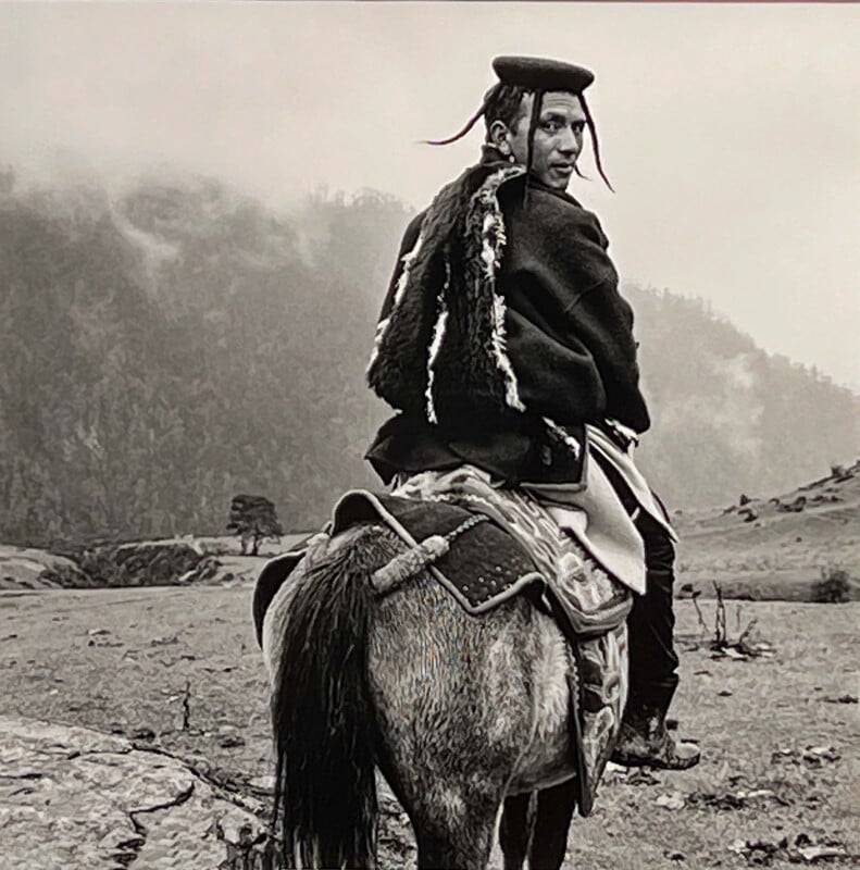 A person wearing traditional clothing and a hat rides a horse across a rugged, misty landscape, looking back over their shoulder with mountains and trees visible in the background.