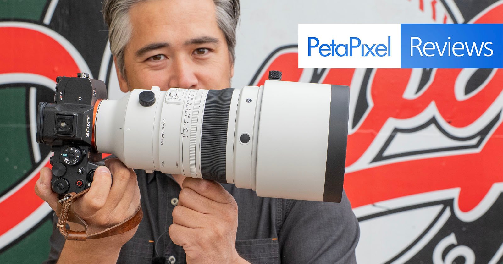 A person holds a professional Sony camera with a large white telephoto lens. The background features a mural and a blue and white “PetaPixel Reviews” banner in the top right corner.