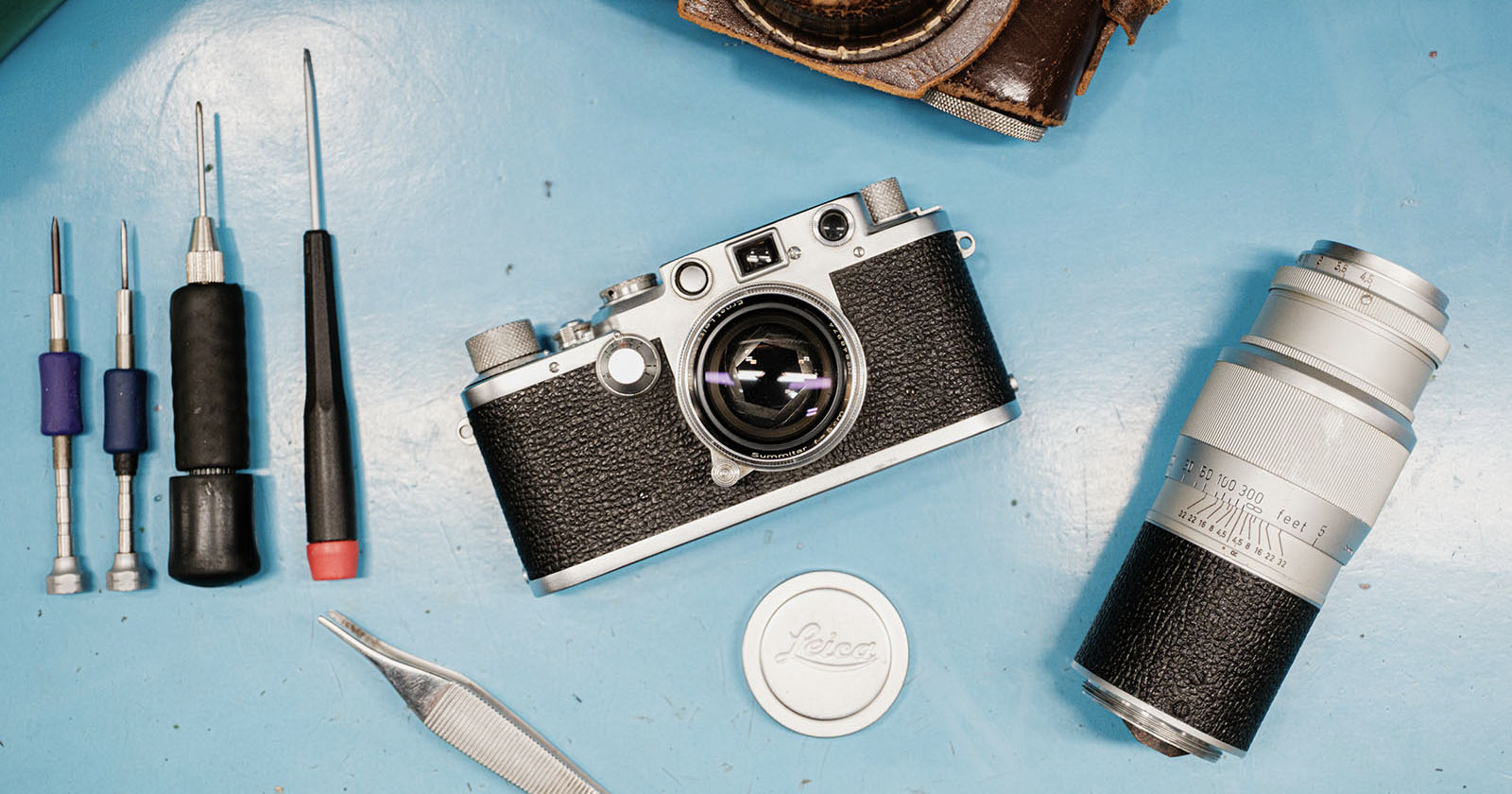 A vintage Leica camera sits on a blue surface surrounded by screwdrivers, tweezers, a camera lens, and a lens cap, suggesting camera repair or maintenance.