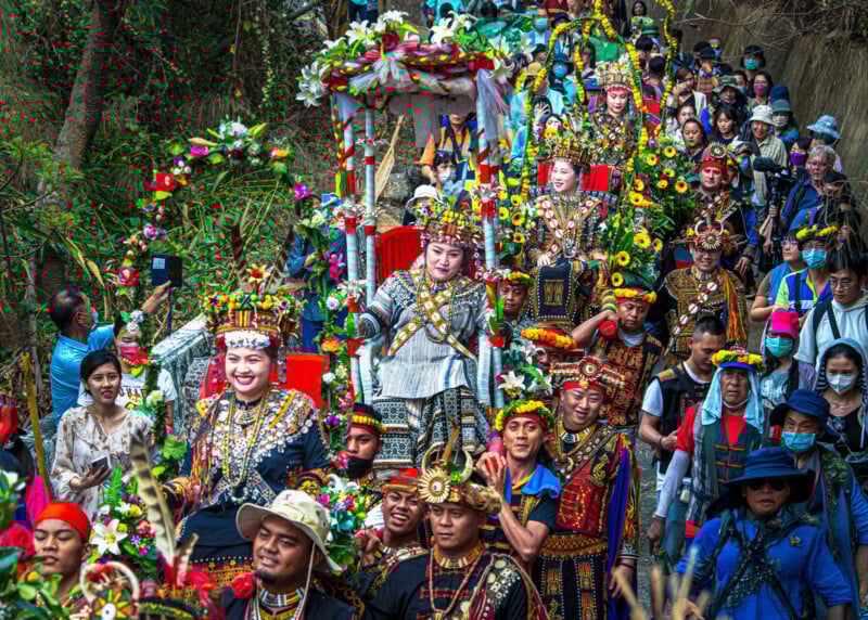 A vibrant parade features people in ornate traditional costumes, colorful headdresses, and masks. They walk through a lush outdoor setting, surrounded by greenery and onlookers, some of whom wear casual clothes and hats.