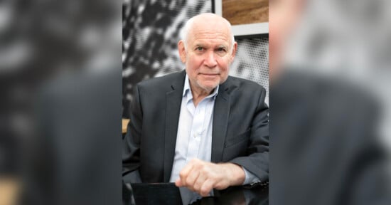 An older man with short white hair, wearing a dark suit jacket and light blue shirt, sits at a table and looks at the camera with a gentle expression. The background is blurred and patterned.