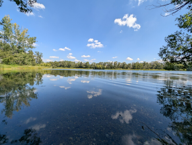 A calm lake with clear water reflecting the blue sky and white clouds, surrounded by green trees under a sunny sky.
