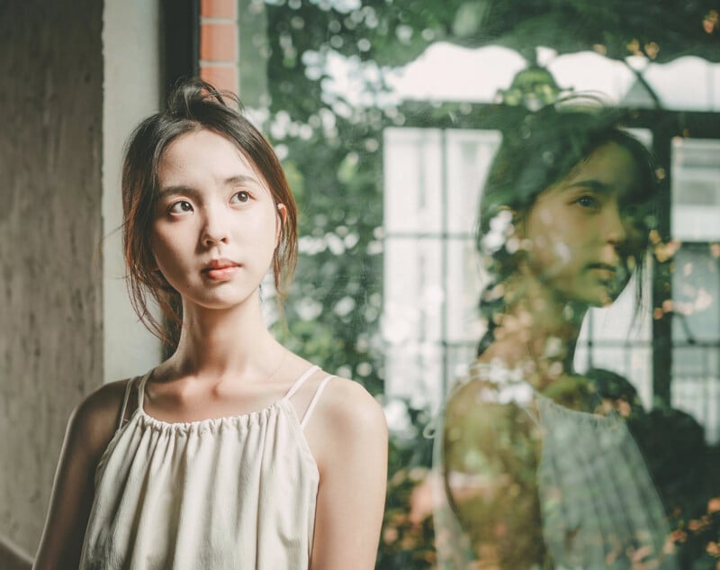 A young woman in a white sleeveless top stands indoors beside a large window, her reflection visible on the glass. She gazes thoughtfully to the side, with soft natural light illuminating her face.