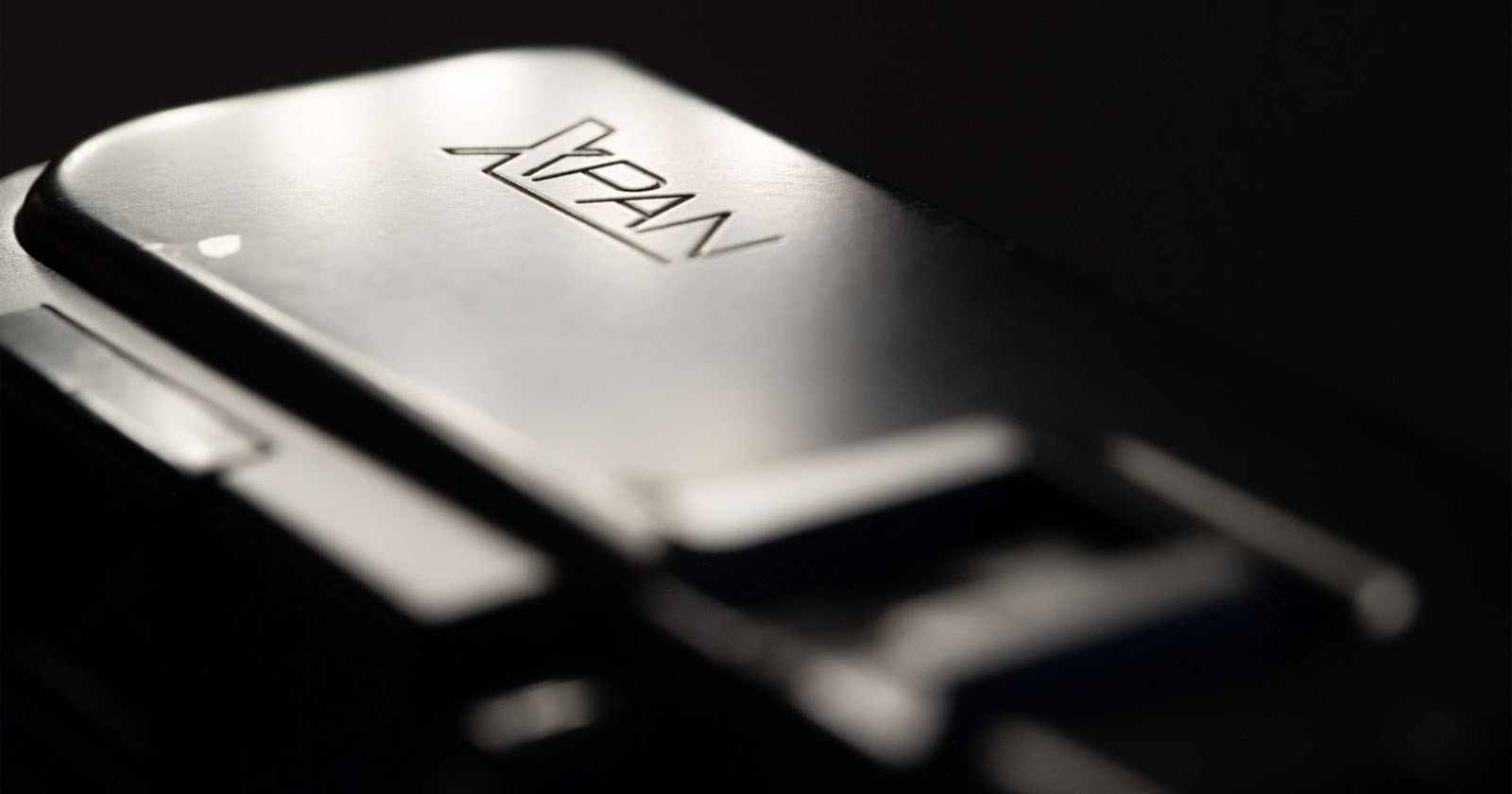 Close-up of a metallic camera part with the engraved logo "XPAN," highlighting its smooth, reflective surface and blurred background.