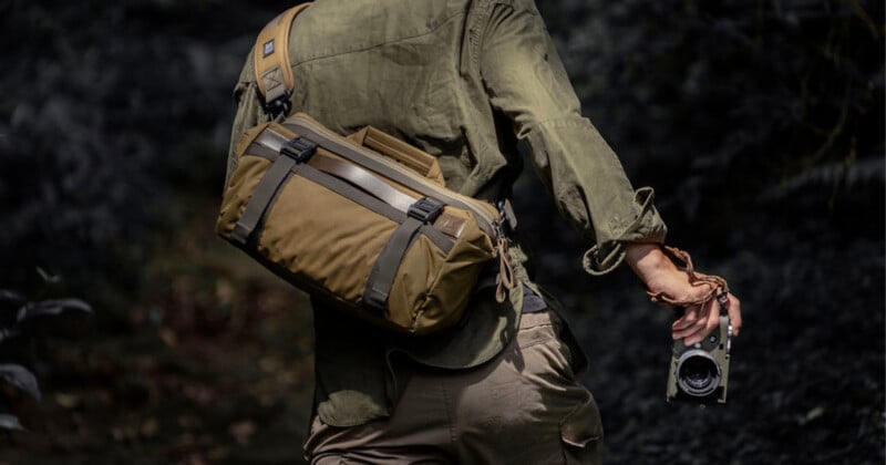 A person wearing olive-green outdoor clothing carries a khaki shoulder bag and holds a camera while walking through a dark, forested area.