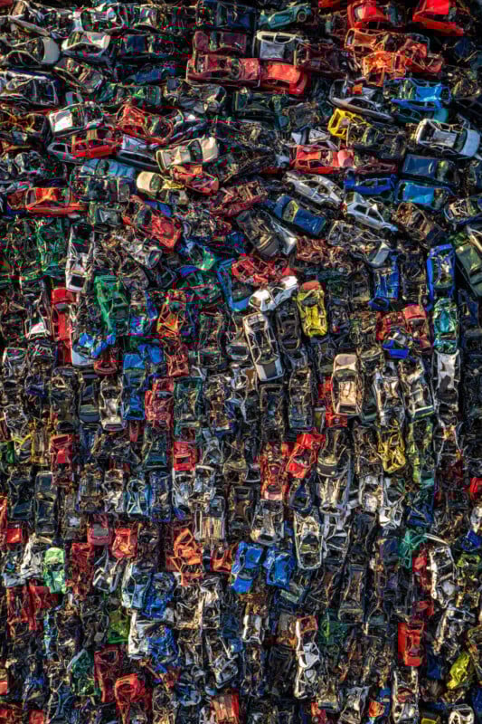 Aerial view of a dense pile of colorful, crushed cars stacked closely together in a junkyard, forming a chaotic, textured pattern.