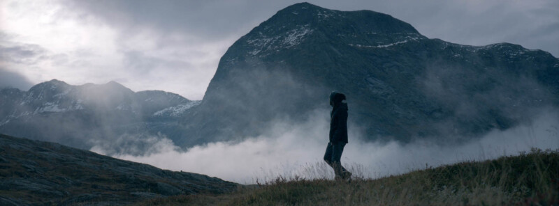 A person stands on a grassy hill in front of a large, misty mountain under a cloudy sky. The scene is serene and dramatic, with low clouds drifting around the mountain peaks.