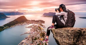 A person with a backpack sits on a rocky cliff, taking photos of a coastal town below, surrounded by water and mountains at sunset. The sky is colorful with shades of pink and orange.
