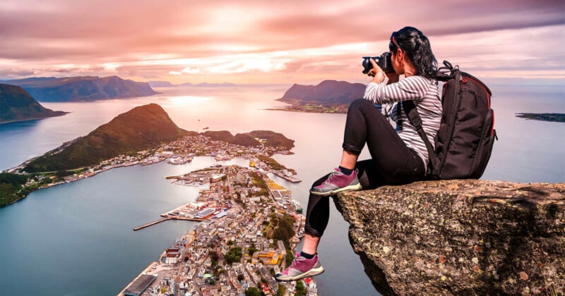 A person with a backpack sits on a rocky cliff, taking photos of a coastal town below, surrounded by water and mountains at sunset. The sky is colorful with shades of pink and orange.