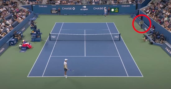 A wide view of a tennis match at the US Open with one player on the court. A person in the stands, circled in red, is leaning over the barrier. Spectators fill the stands, and the scoreboard is visible.
