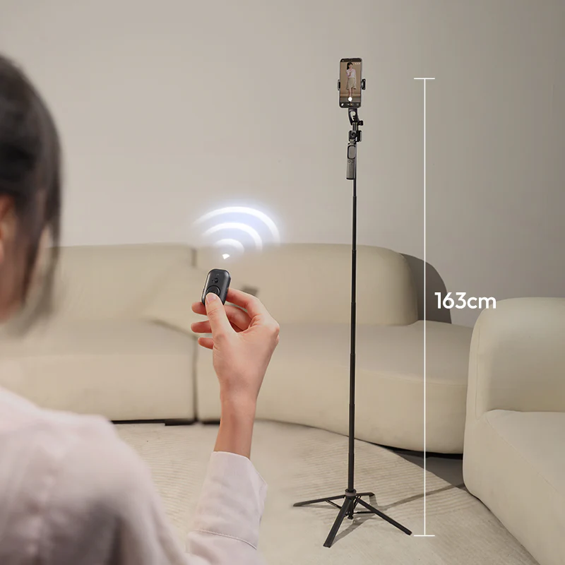 A person holds a remote control facing a smartphone mounted on a 163cm tall tripod in a living room with a beige sofa in the background.