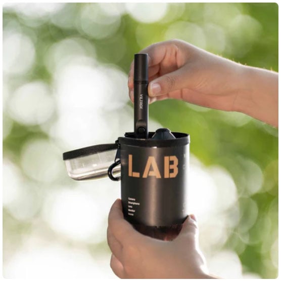 A person’s hands placing a VLAB vaporizer pen into a black container labeled “LAB,” with a blurred green outdoor background.