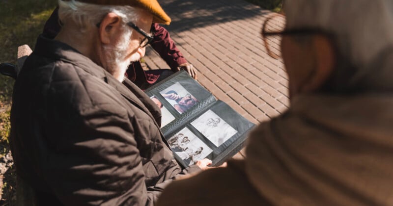 Two elderly people sit on a bench outdoors, looking at a photo album filled with black-and-white pictures. Sunlight casts shadows on the wooden path beside them.