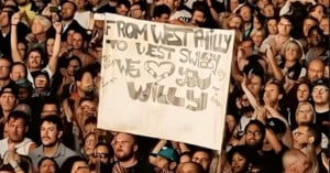 A large crowd at a concert holds a handmade sign reading, “FROM WEST PHILLY TO WEST SWIDAY WE [heart] YOU WILLY!” People around the sign are cheering and clapping.