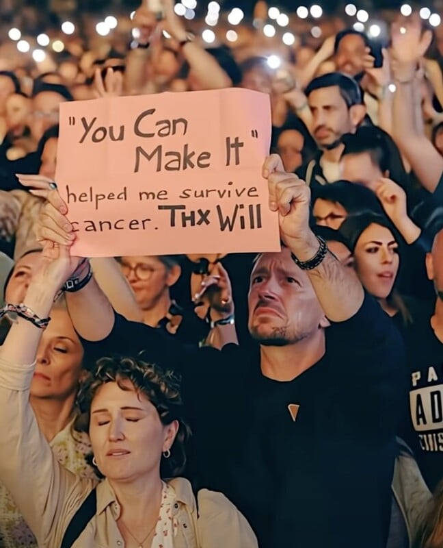 A man in a crowd holds up a pink sign reading, "You Can Make It," helped me survive cancer. Thx Will." People around him look emotional, and many hold up their phones with flashlights on.