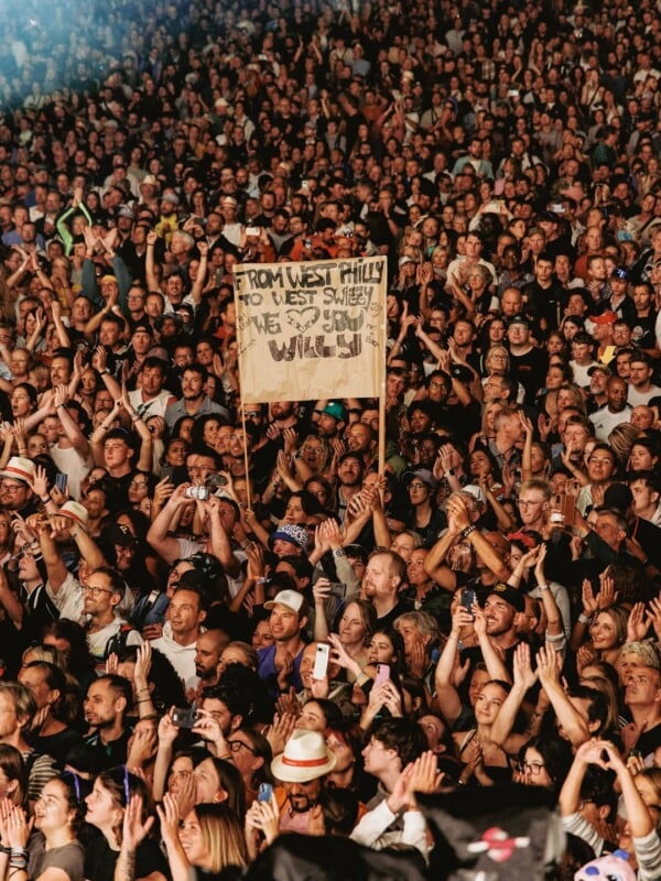 A large crowd at a concert, many holding up phones. In the center, a group lifts a cardboard sign reading, “FROM WEST PHILLY TO WEST SWITZ WE LOVE YOU WILL!!!” The crowd looks excited and engaged.