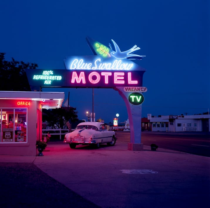 A vintage neon sign reading "Blue Swallow Motel" glows at dusk, with an old-fashioned car parked beneath it. The motel office is lit with pink and blue lights, evoking a retro vibe along a quiet roadside.