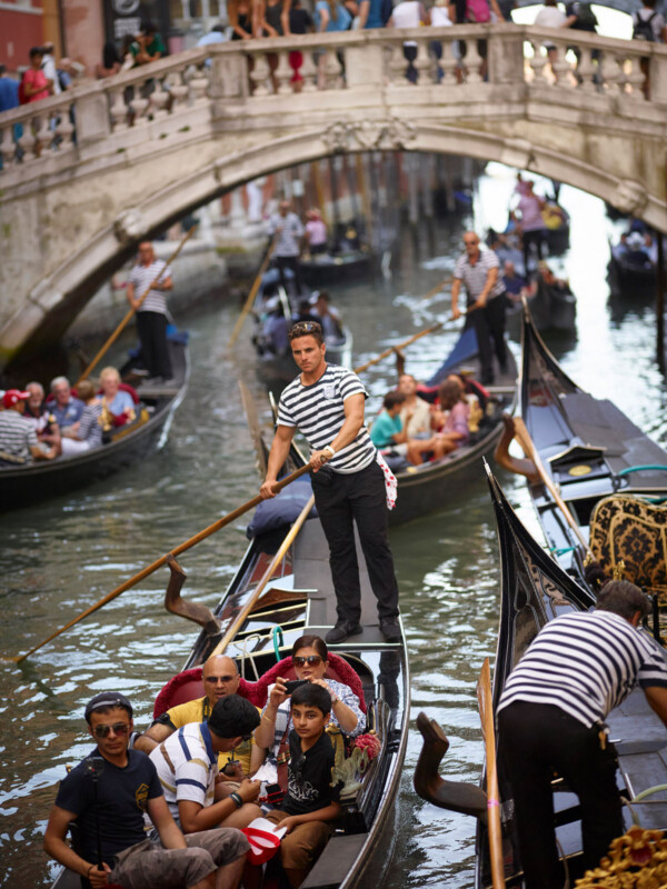 Gondoliers in striped shirts steer gondolas filled with tourists along a crowded canal in Venice, Italy, with a stone bridge and people watching from above in the background.