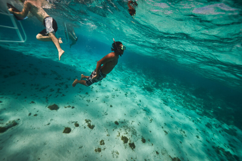 Two people snorkel underwater in clear blue ocean near a boat. Sunlight filters through the water, illuminating the sandy sea floor below with patches of seaweed.