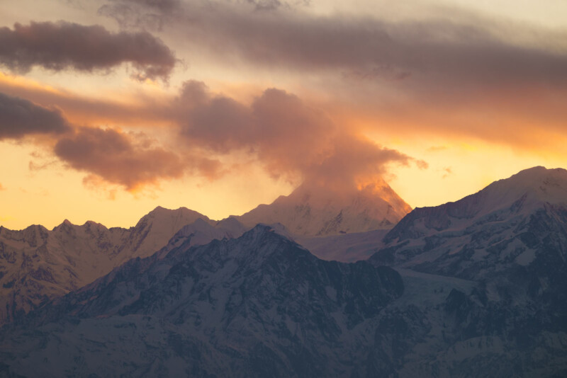 Snow-covered mountain peaks are silhouetted against a dramatic sunset sky with vibrant orange and yellow hues, while clouds partially obscure the tallest peak.