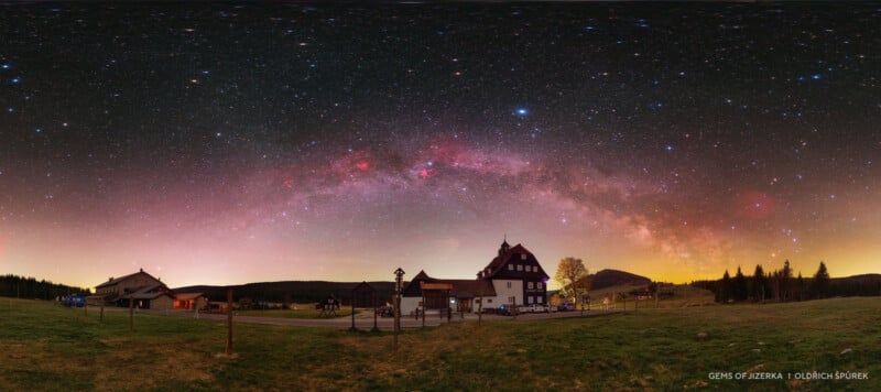 A panoramic night sky with the Milky Way arching over a rural landscape, including a large house, smaller buildings, grassy fields, and distant hills under a star-filled sky.