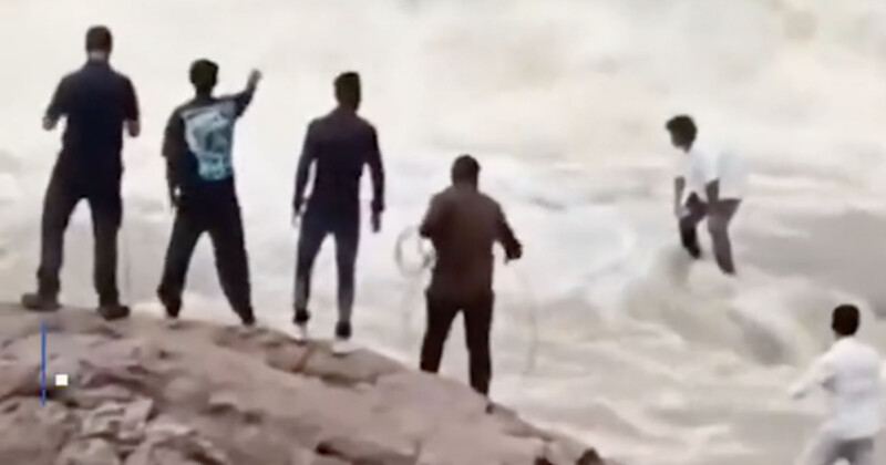 Cinco personas se pararon en el banco rocoso, algunas sosteniendo cuerdas mientras una en el agua. La escena se ve tensa, lo que indica que es probable que un intento de rescate esté en marcha. El agua es agitada y se mueve rápidamente.