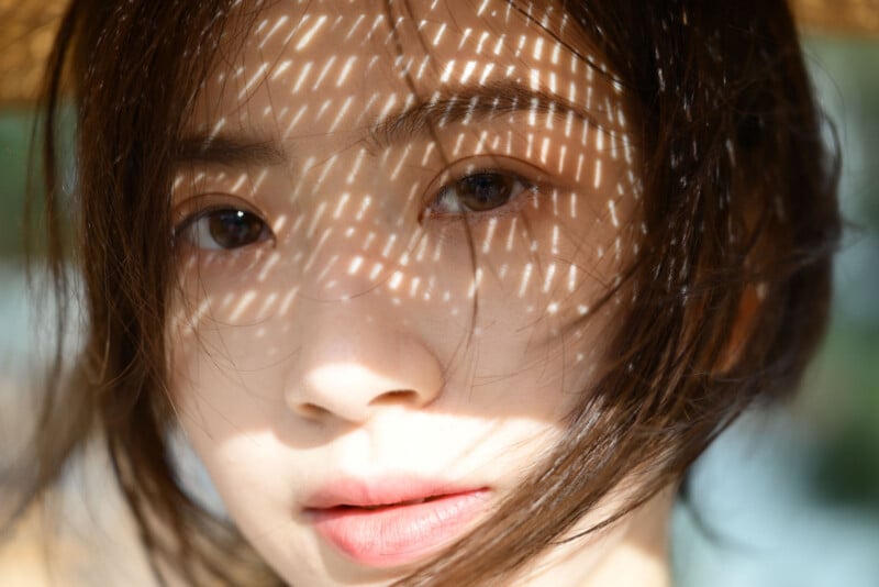 Close-up of a person’s face with short dark hair, softly lit. Dappled light patterns from a woven object create bright spots and shadows across their skin. Their expression is calm and thoughtful.