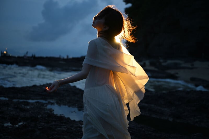 A person in a flowing white dress stands on rocky shore at dusk, backlit by warm light that illuminates their hair and dress, with the ocean and dark cliffs in the background.