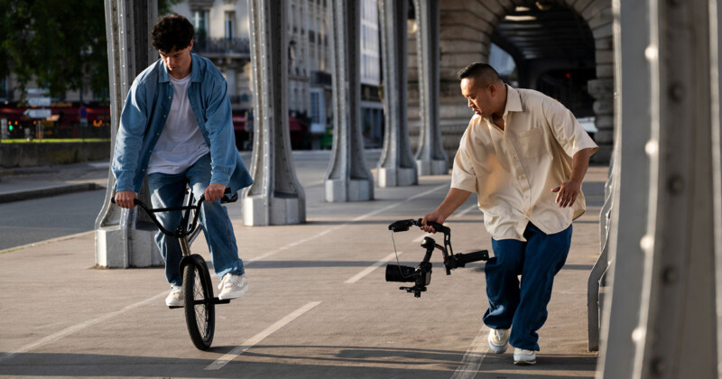A young man rides a BMX bike on a city bridge while another man walks beside him, filming with a professional camera rig. The scene takes place during the day under metal arches.
