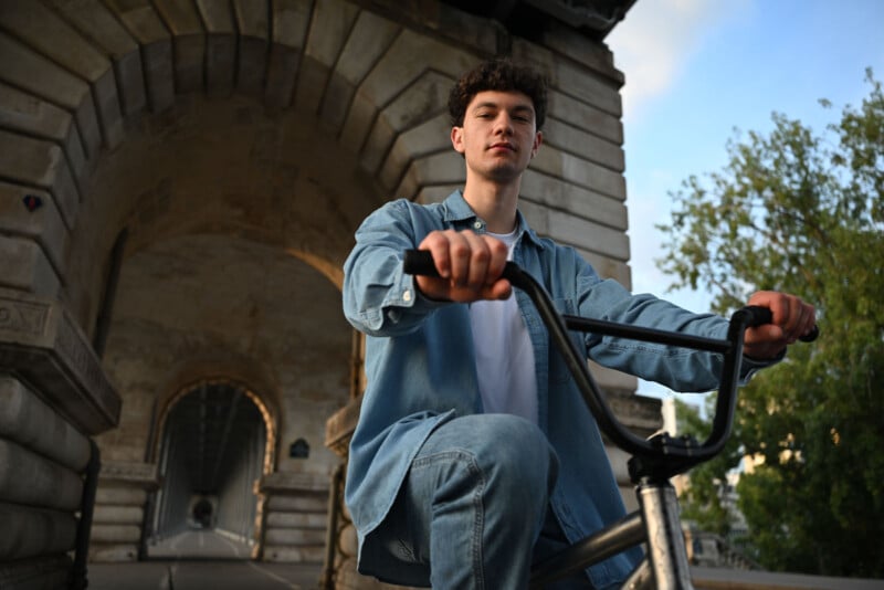 A young man in a denim jacket and jeans sits on a bicycle, looking at the camera, with a stone archway and trees in the background.