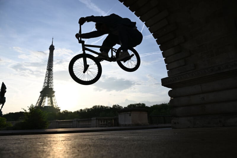 A person on a BMX bike jumps in silhouette under an archway, with the Eiffel Tower visible in the background at sunset.