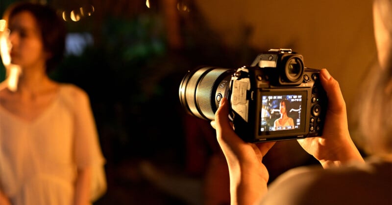 A person holds a camera and takes a photo of a woman in a light dress, who stands softly lit in the background; the woman's image is clearly visible on the camera screen.