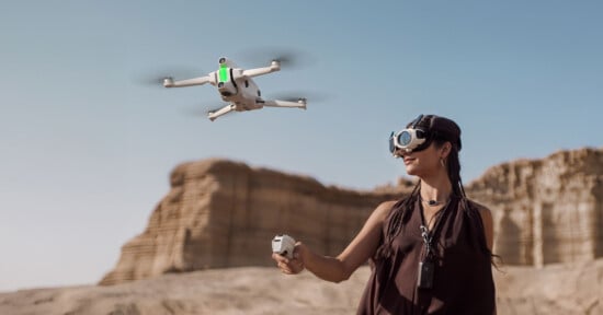 A person wearing virtual reality goggles flies a white drone using a remote controller in a desert landscape with rock formations in the background under a clear blue sky.