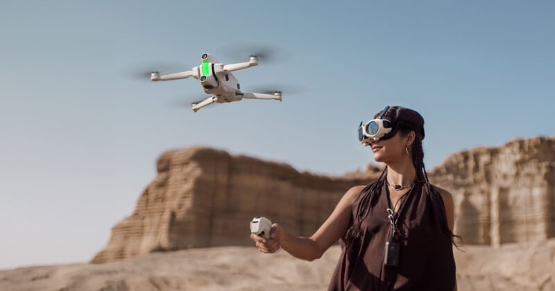 A person wearing virtual reality goggles flies a white drone using a remote controller in a desert landscape with rock formations in the background under a clear blue sky.