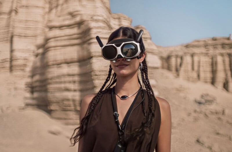 A woman with braided hair stands in a rocky desert landscape, wearing futuristic, oversized VR goggles with antennae. She is dressed in a sleeveless dark top and a pendant necklace.