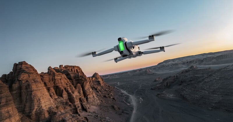 A white drone with green lights flies over a rugged, rocky landscape at sunset, with a clear sky and dramatic cliffs in the foreground.