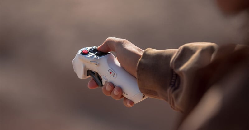A person wearing a brown jacket holds a white handheld electronic controller with buttons and a joystick, against a blurred outdoor background.
