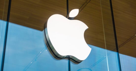 A large white Apple logo is mounted on the glass exterior of an Apple Store, reflecting the sky and wooden ceiling inside.