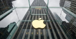 A large, glowing Apple logo is displayed inside a glass cube at the Apple Store on Fifth Avenue in New York City, with tall skyscrapers and cloudy skies reflected in the glass.