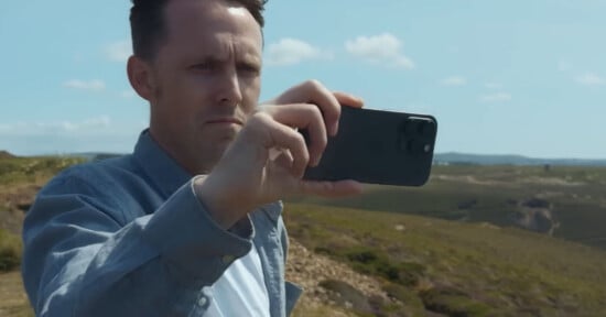 A man standing outdoors on a hilltop uses a smartphone to take a photo or video, with a landscape of rolling hills and a partly cloudy sky in the background.