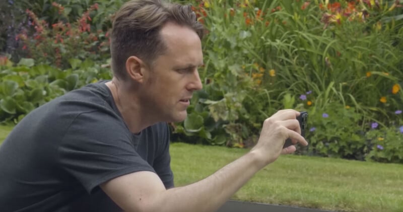 A man in a dark t-shirt kneels on the grass in a garden, holding a small camera or device and focusing intently, with colorful flowers and greenery in the background.