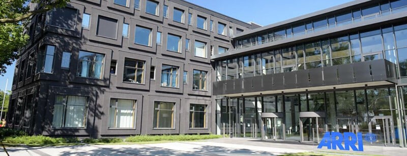 A modern office building with large windows and a glass entrance. The ARRI logo is visible on the ground near the entrance. Trees and grass are in the foreground under a clear blue sky.