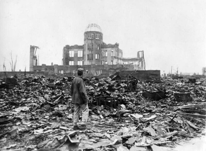 A person stands amid widespread rubble and debris, facing the ruined Hiroshima Prefectural Industrial Promotion Hall, heavily damaged after the atomic bombing in 1945. The dome structure is partially intact.
